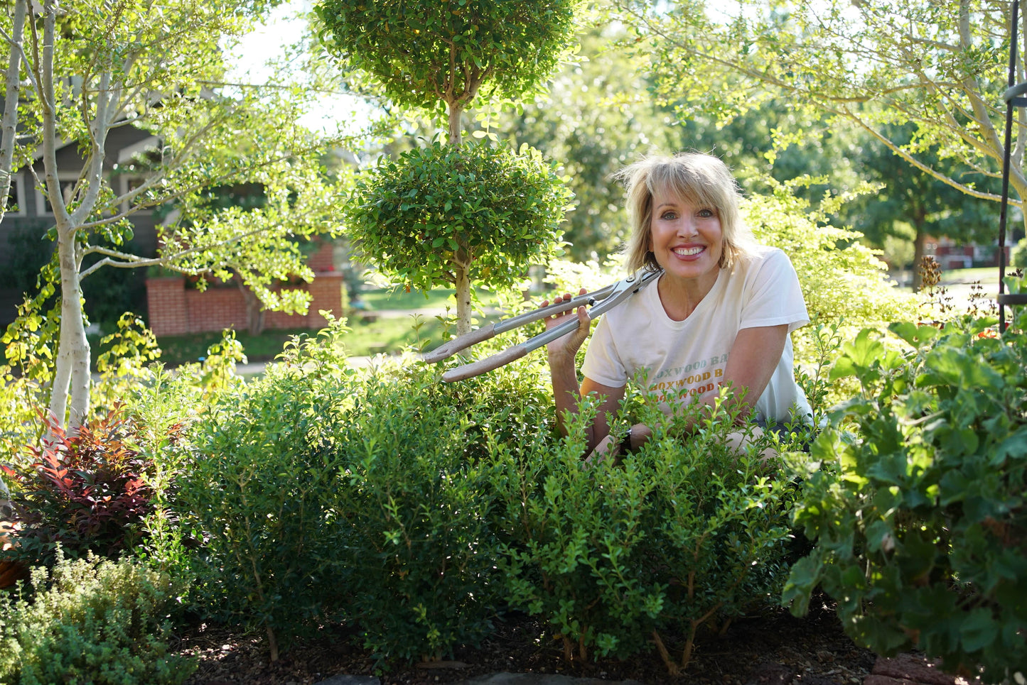 Linda Vater in a white t-shirt kneels in a garden holding pruning shears, surrounded by green bushes and trees on a sunny day.