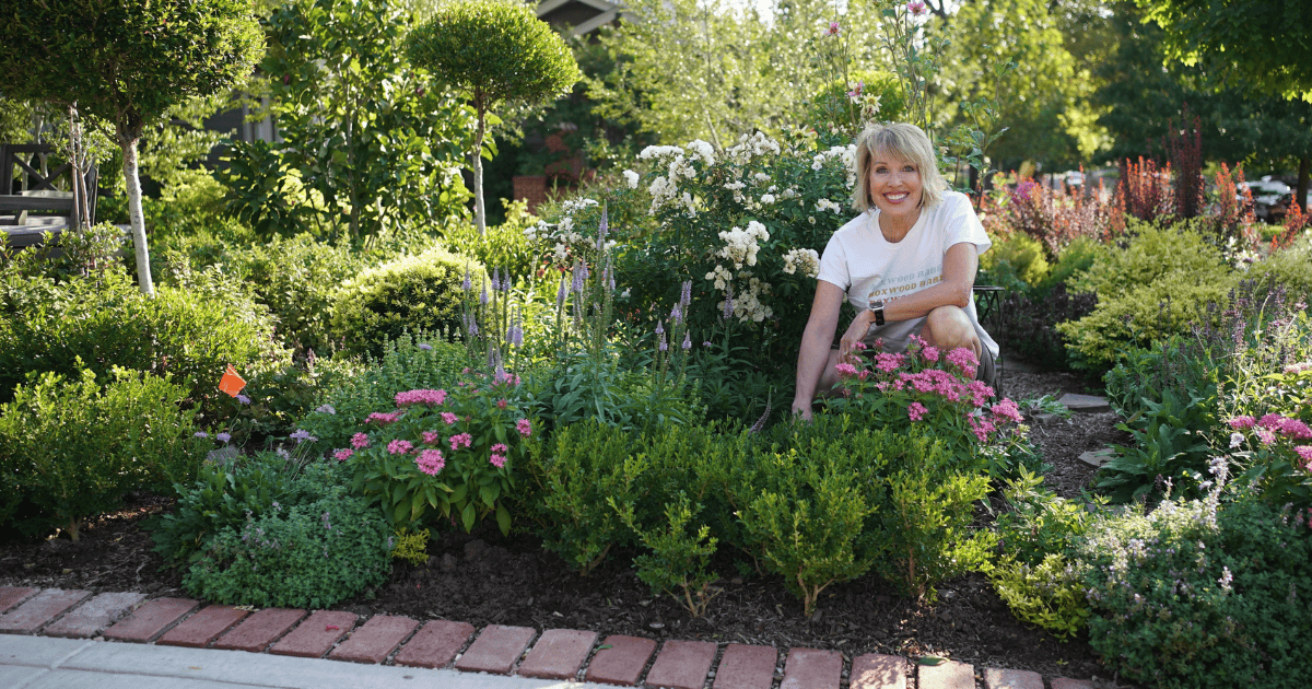 A person kneels in a lush garden surrounded by various flowers and greenery, smiling at the camera. Brick edging defines the garden path.