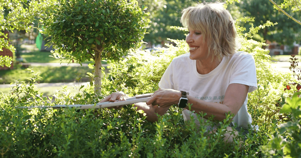 A woman with short blonde hair tends to a garden, smiling as she trims a bush. She wears a white T-shirt and a watch, surrounded by lush greenery and sunlight.