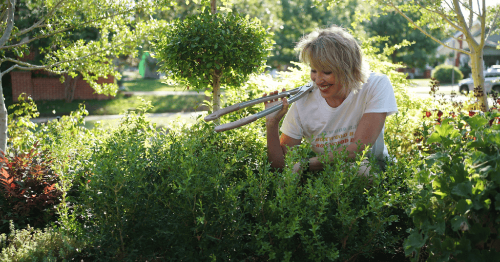 A person smiles while trimming bushes in a garden using a pair of hedge shears on a sunny day.