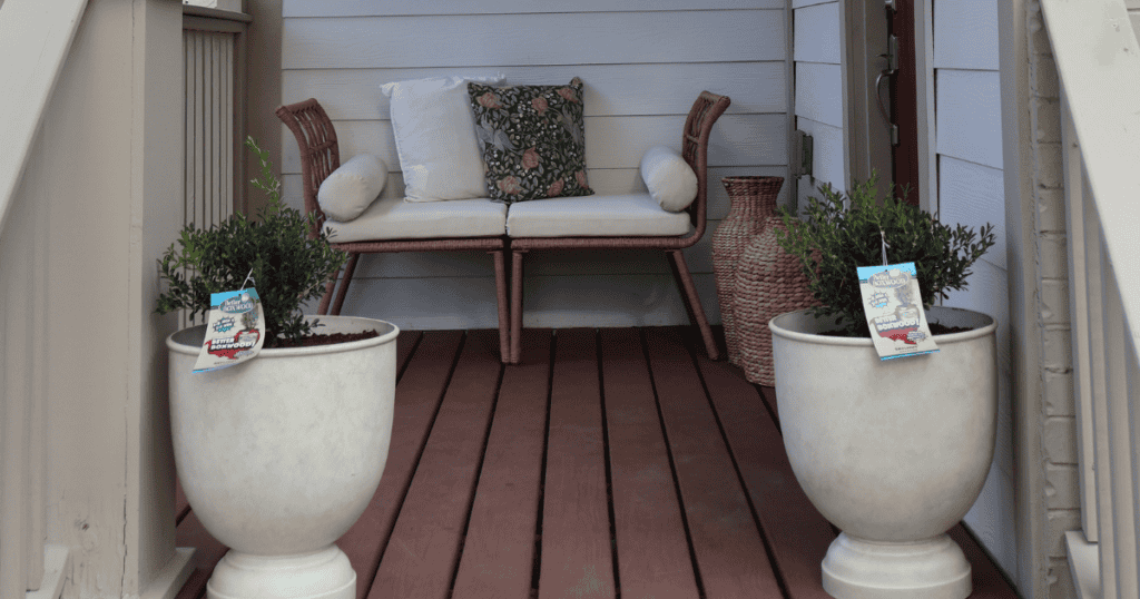 A porch with a wicker loveseat, two white cushions, a patterned pillow, and two potted plants on either side. A wicker basket is next to the seat.